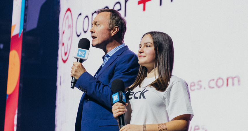 Two people holding microphones on stage at an event, with a logo in the background.